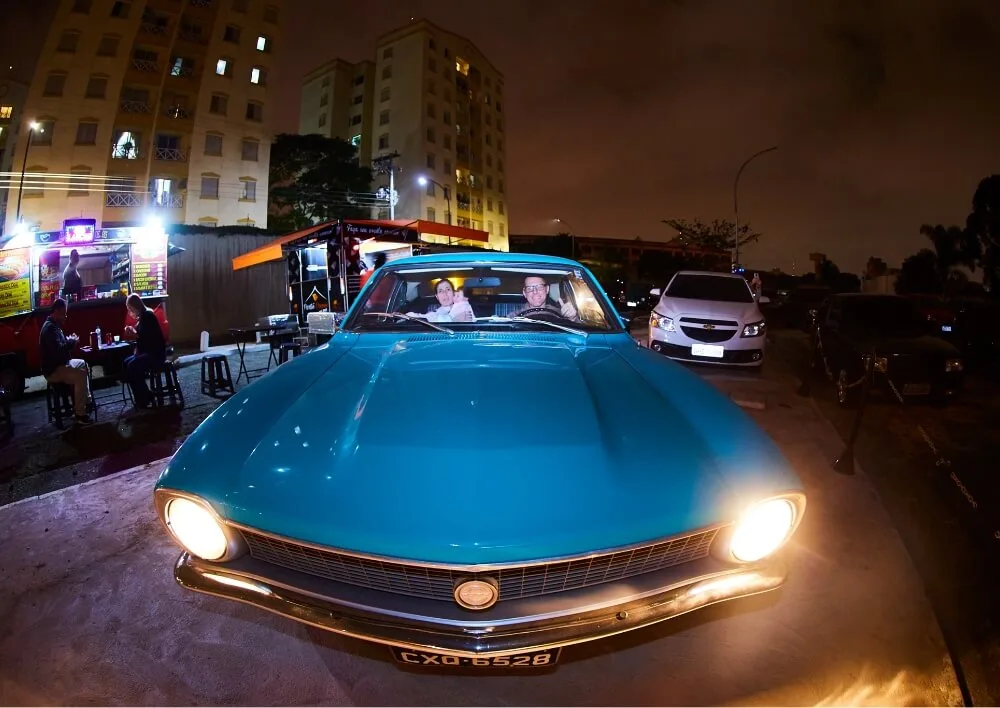 Casal sorrindo dentro de um carro clássico azul com os faróis acesos